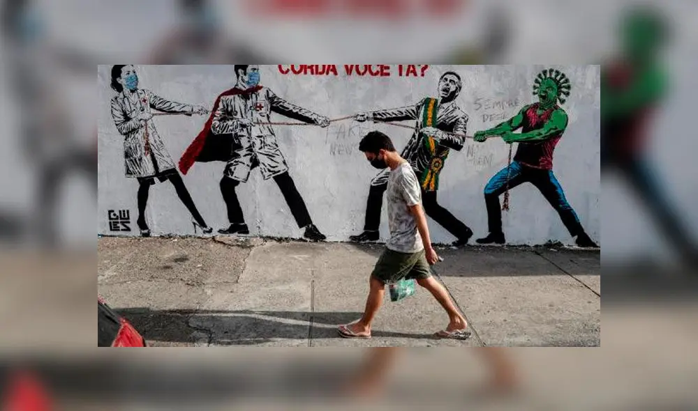 Un hombre pasa al frente de un graffiti que representa al presidente brasileño Jair Bolsonaro y una figura que representa el nuevo coronavirus COVID-19 tirando de una cuerda contra los trabajadores de la salud. Foto: EFE. Un hombre pasa al frente de un graffiti que representa al presidente brasileño Jair Bolsonaro y una figura que representa el nuevo coronavirus COVID-19 tirando de una cuerda contra los trabajadores de la salud. Foto: EFE.