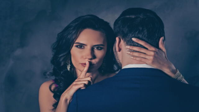 Shh! Portrait of tempting brunette lady showing silence sign with forefinger touching secret mysterious gentlemen with rear view, lovely Mr and Mrs isolated on grey background (Shh! Portrait of tempting brunette lady showing silence sign with forefing