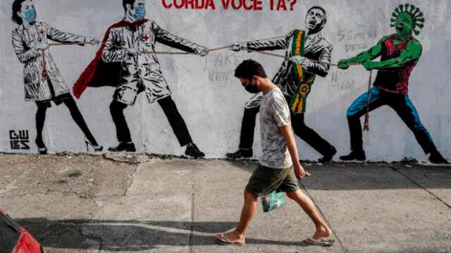Un hombre pasa al frente de un graffiti que representa al presidente brasileño Jair Bolsonaro y una figura que representa el nuevo coronavirus COVID-19 tirando de una cuerda contra los trabajadores de la salud. Foto: EFE.