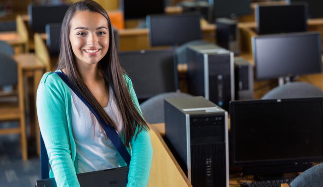Teenage Hispanic girl is student in public high school. She is looking at the camera and smiling while walking in high school computer lab classroom or library. She is wearing a messenger bag style backpack over casual layered clothing. Student is holding a laptop computer. She is near rows of desks with desktop computers.