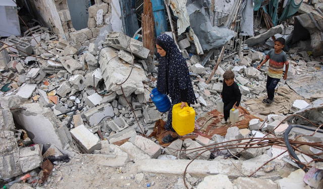 Una mujer palestina desplazada y varios niños caminan sobre los escombros de un edificio mientras transportan contenedores de agua en el barrio de Shujaiya. Foto: AFP   